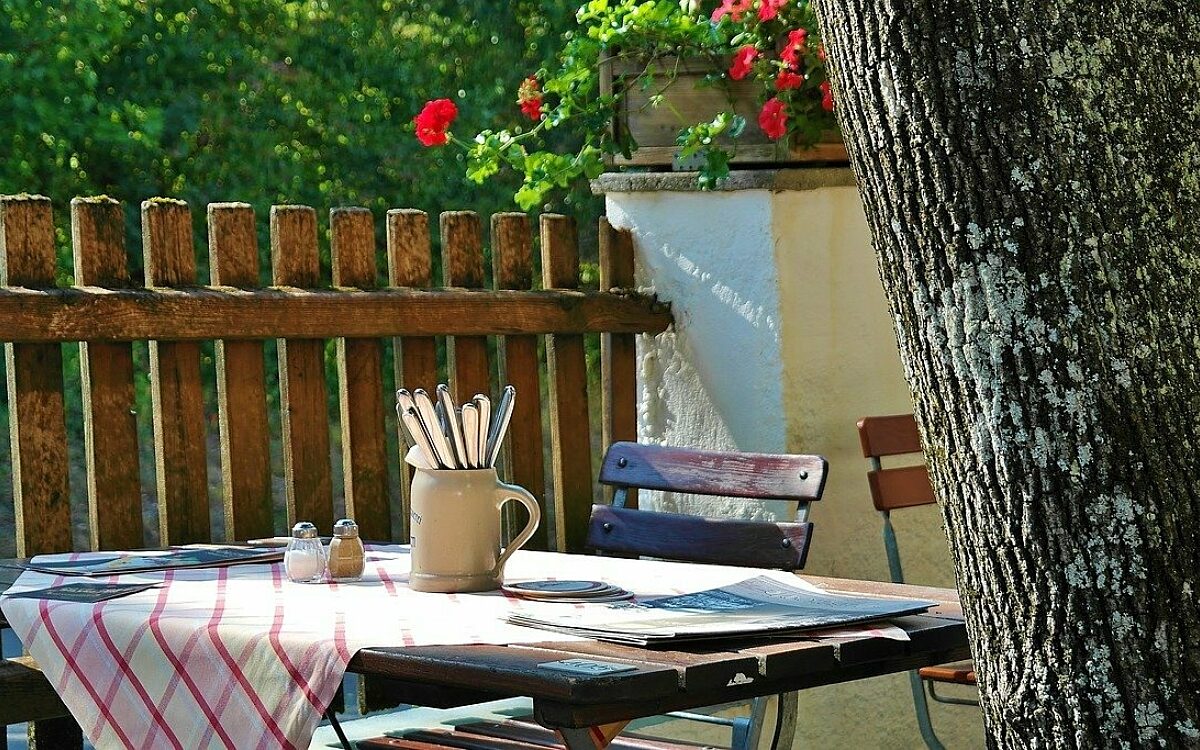 In einem Biergarten steht ein eingedeckter Tisch hinter einem Baum. In einem Biergarten steht ein eingedeckter Tisch hinter einem Baum.